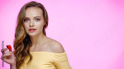 Young woman holding strawberry while looking at camera on pink