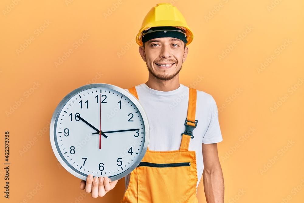 Hispanic young man wearing builder uniform and hardhat holding clock looking positive and happy standing and smiling with a confident smile showing teeth Hispanic young man wearing builder uniform and hardhat holding clock looking positive and happy standing and smiling with a confident smile showing teeth