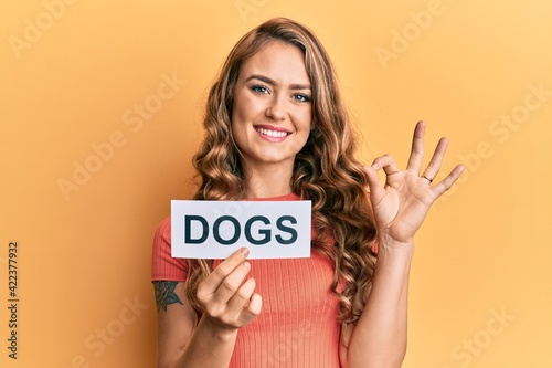 Young blonde girl holding paper with dogs message doing ok sign with fingers, smiling friendly gesturing excellent symbol