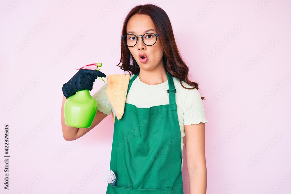 Young beautiful chinese girl wearing apron holding sprayer scared and amazed with open mouth for surprise, disbelief face Young beautiful chinese girl wearing apron holding sprayer scared and amazed with open mouth for surprise, disbelief face