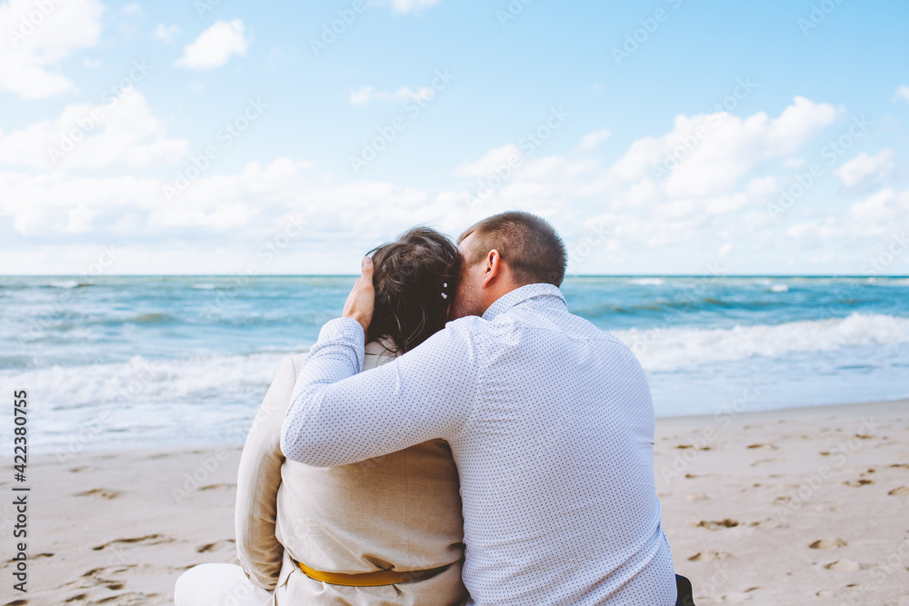 Back view of happy just married middle age couple seat on big stone at beach against blue sky with clouds