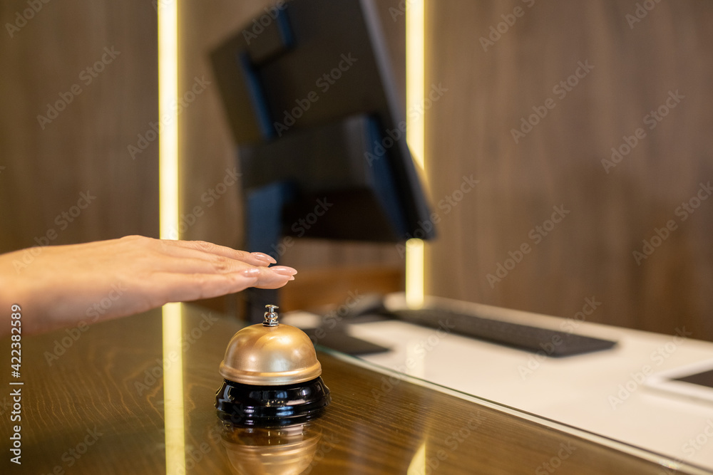 Hand of young woman over bell on wooden reception counter going to push button and call for receptionist to registrate her in hotel room Hand of young woman over bell on wooden reception counter going to push button and call for receptionist to registrate her in hotel room