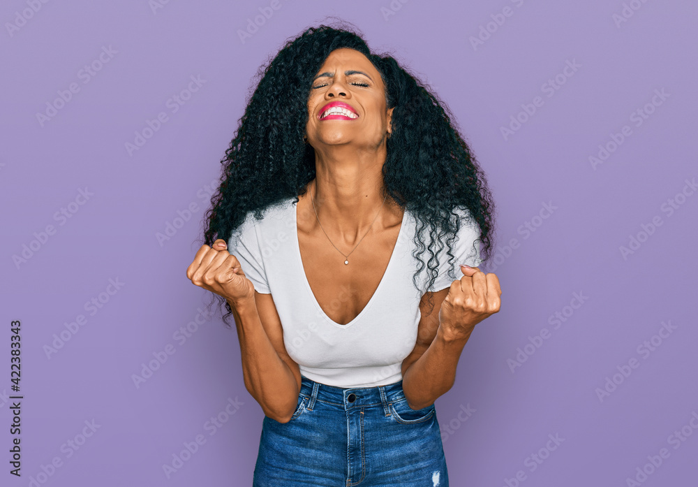 Middle age african american woman wearing casual white t shirt very happy and excited doing winner gesture with arms raised, smiling and screaming for success. celebration concept. Middle age african american woman wearing casual white t shirt very happy and excited doing winner gesture with arms raised, smiling and screaming for success. celebration concept.