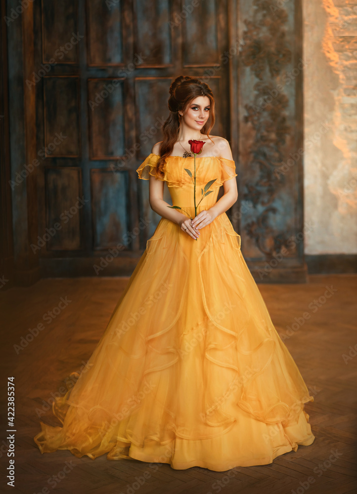 Girl beauty fantasy princess in yellow long historical, medieval silk dress holding flower red rose in her hands. Background of old gothic castle room. Fairy tale bewitched queen. Happy woman smiles