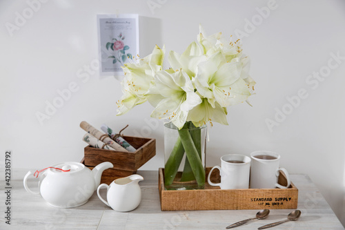 A bouquet of white lily in a glass vase on a table with two tall cups of coffee, a teapot, spoons, and a milk jug.