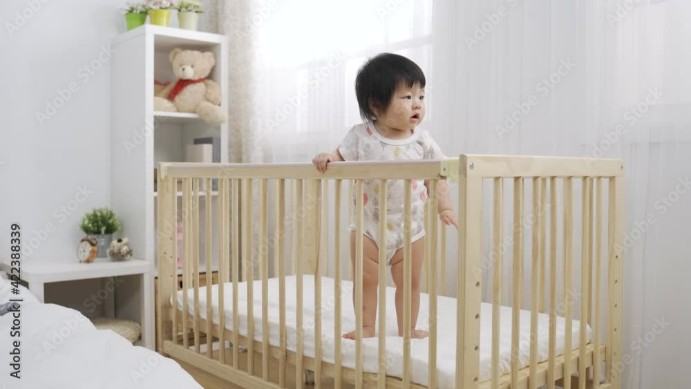 full length innocent toddler girl standing in the baby crib is staring into the space anticipating her parents to come over at a cozy bedroom interior. full length innocent toddler girl standing in the baby crib is staring into the space anticipating her parents to come over at a cozy bedroom interior.
