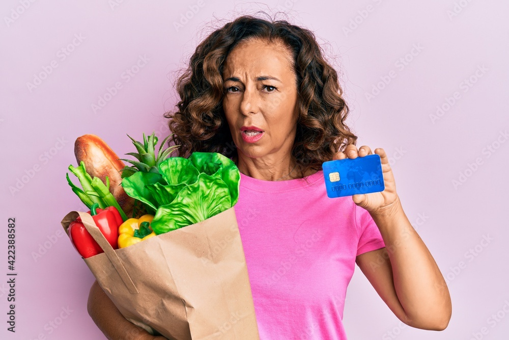 Middle age hispanic woman holding groceries and credit card clueless and confused expression. doubt concept. Middle age hispanic woman holding groceries and credit card clueless and confused expression. doubt concept.