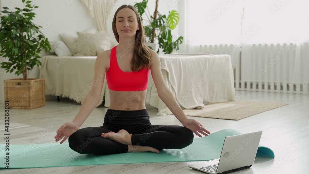 Positive yoga woman does practice in front of a laptop while sitting on a sports mat at home Positive yoga woman does practice in front of a laptop while sitting on a sports mat at home