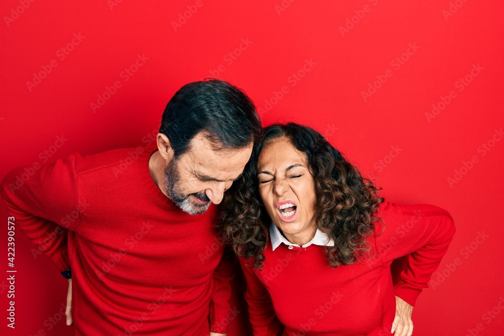 Middle age couple of hispanic woman and man hugging and standing together suffering of backache, touching back with hand, muscular pain Middle age couple of hispanic woman and man hugging and standing together suffering of backache, touching back with hand, muscular pain
