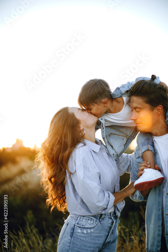 happy young family in the park at sunset. happy boy sits on dad's shoulders. mom kisses her son. sun rays on faces. young stylish family in casual outdoor clothes 