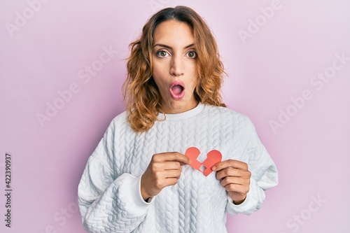 Young caucasian woman holding broken heart paper shape afraid and shocked with surprise and amazed expression, fear and excited face.