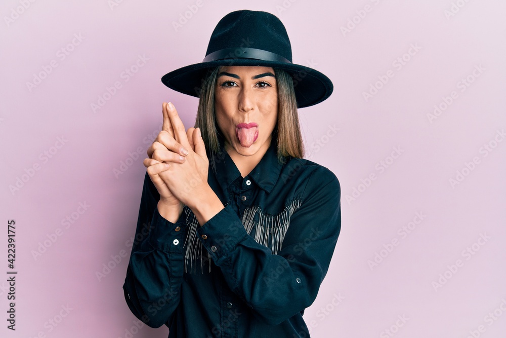 Young hispanic woman wearing cowgirl style doing pistol gesture with hands sticking tongue out happy with funny expression. Young hispanic woman wearing cowgirl style doing pistol gesture with hands sticking tongue out happy with funny expression.