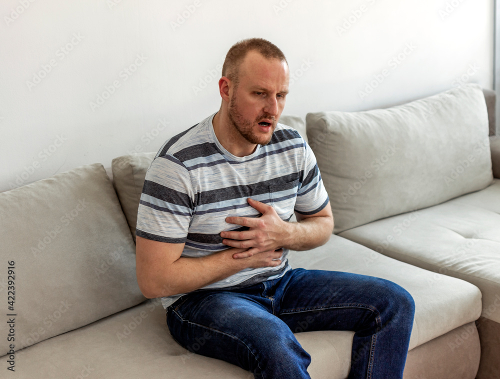 Shot of young man with stomach ache sitting on the bed at home. Man suffering from abdominal pain while sitting on bed at home. Young man having stomach ache while spending time at home alone.