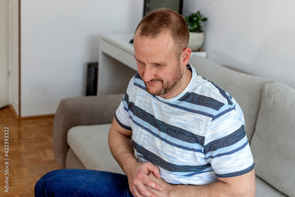 Young man suffering from stomach ache, sitting on a sofa in the living room. Man holding stomach in pain with stomach ache or indigestion. Male holding stomach suffering from pain or diarrhea problem.