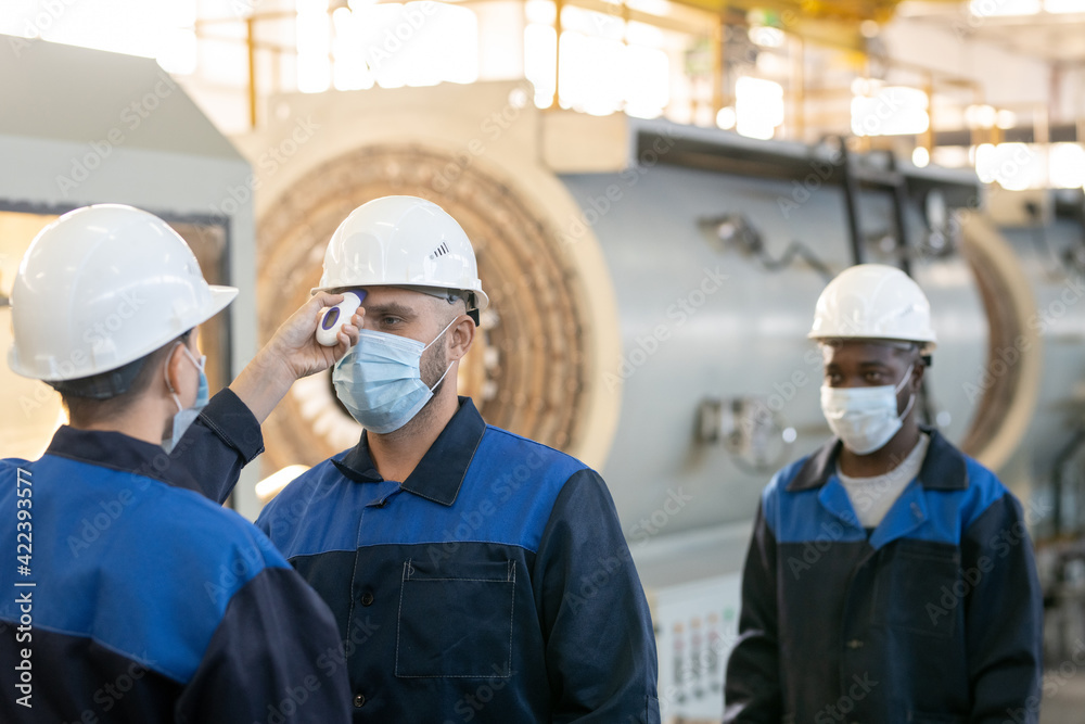 One of several factory workers with thermometer measuring body temperature of his colleagues or subordinates inside large workshop One of several factory workers with thermometer measuring body temperature of his colleagues or subordinates inside large workshop