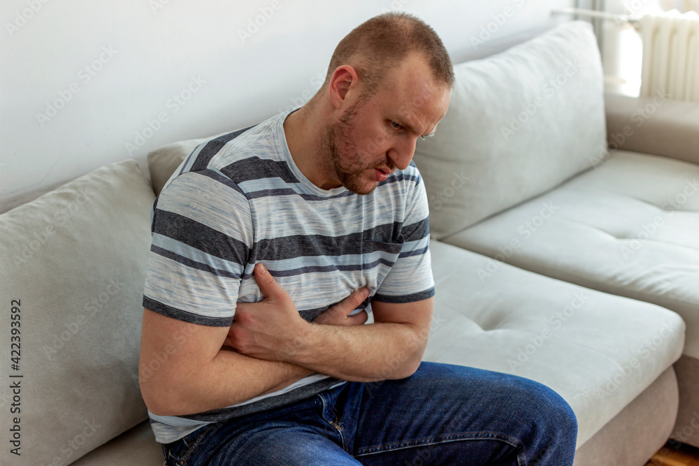 The man is sitting on a beige couch and holding his belly. The man suffers from stomach ache, gastric problems. Abdominal pain, suffering and pain. Medicine and health concept. The man is sitting on a beige couch and holding his belly. The man suffers from stomach ache, gastric problems. Abdominal pain, suffering and pain. Medicine and health concept.