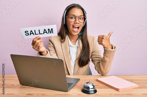 Beautiful hispanic woman wearing operator headset showing sallam greeting pointing thumb up to the side smiling happy with open mouth photo