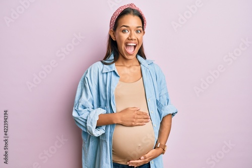 Beautiful hispanic woman expecting a baby, touching pregnant belly celebrating crazy and amazed for success with open eyes screaming excited. photo