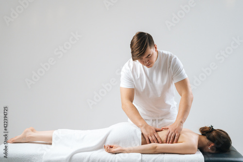 Side view of male masseur with strong hands massaging back of young female lying on massage table on white background. Beautiful skinny naked young woman with perfect skin getting relaxing massage.