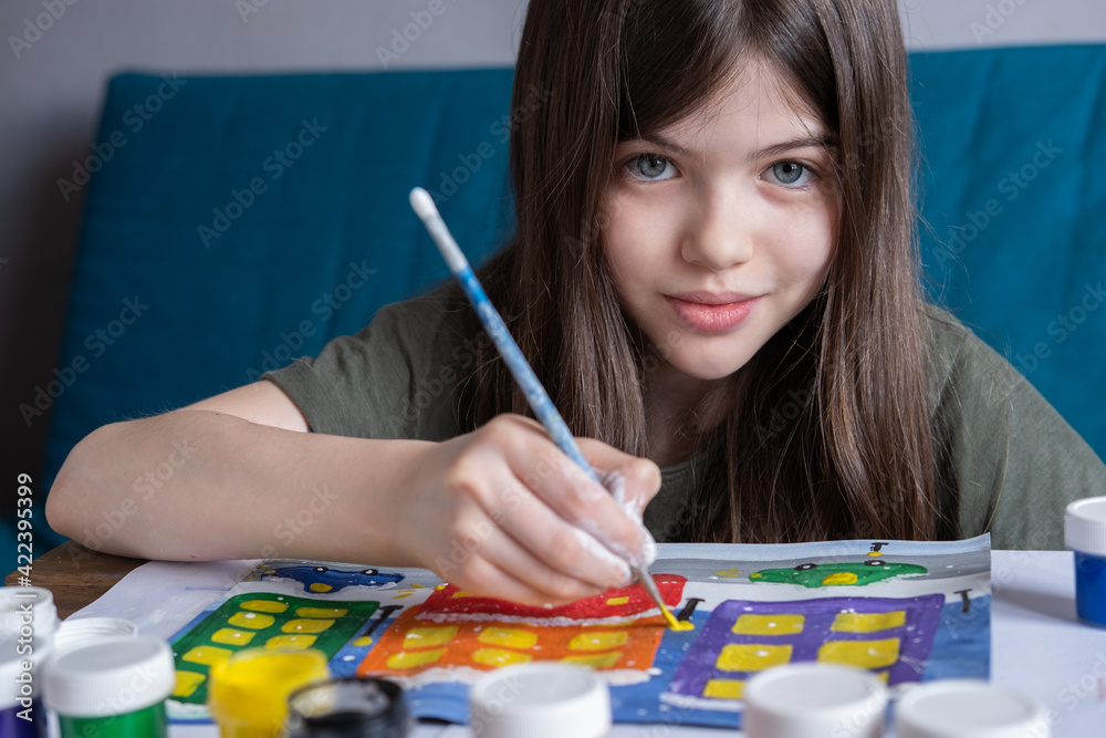 A small, young girl holds a gouache drawing of a winter city in her hands. Children's creativity concept