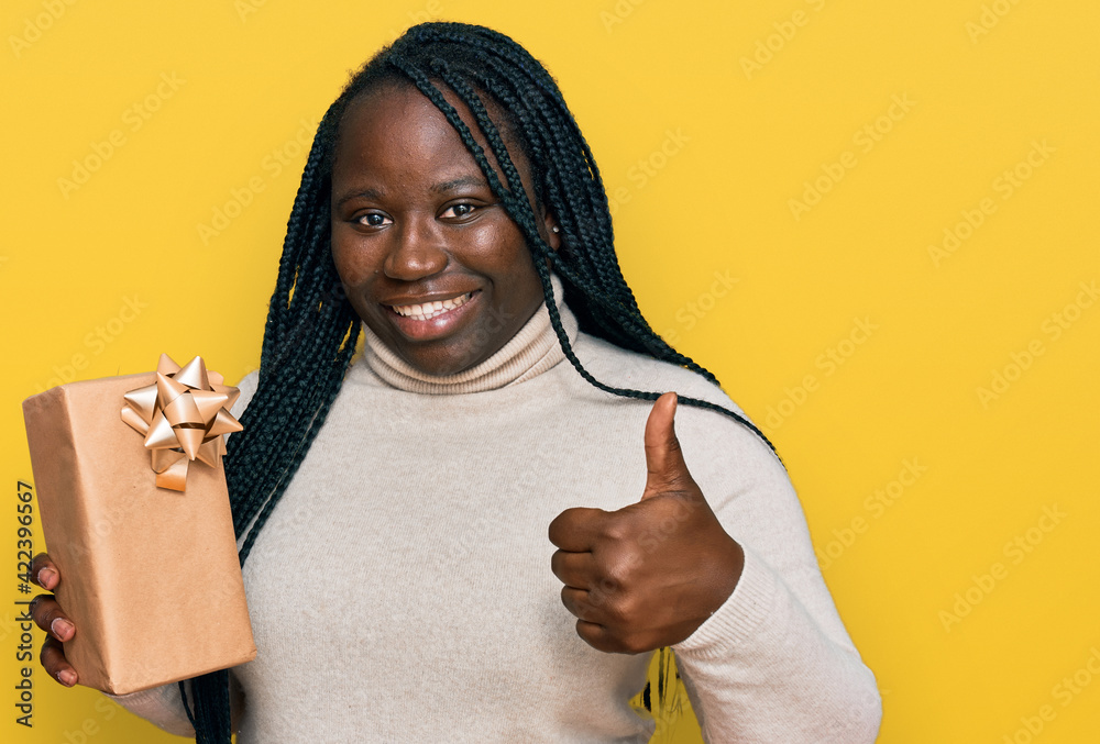 Young black woman with braids holding gift smiling happy and positive, thumb up doing excellent and approval sign Young black woman with braids holding gift smiling happy and positive, thumb up doing excellent and approval sign