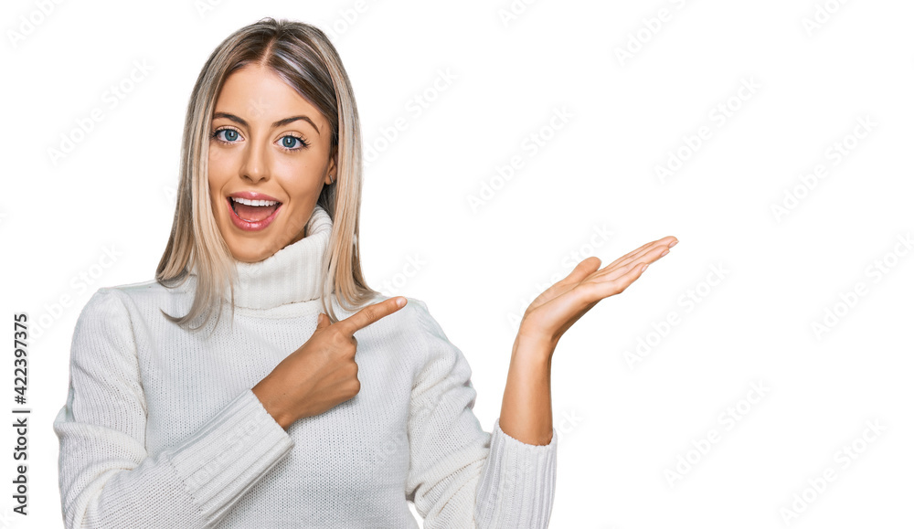 Beautiful blonde woman wearing casual turtleneck sweater amazed and smiling to the camera while presenting with hand and pointing with finger. Beautiful blonde woman wearing casual turtleneck sweater amazed and smiling to the camera while presenting with hand and pointing with finger.