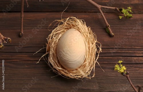 Wooden egg in a nest made of hay and a branch with leaves and buds on a dark natural background. Spring Easter holiday greeting card. Happy Easter concept in eco style.