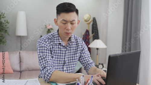 male employee is referring to the document as he is preparing a budge plan for his company on the laptop while working from home.