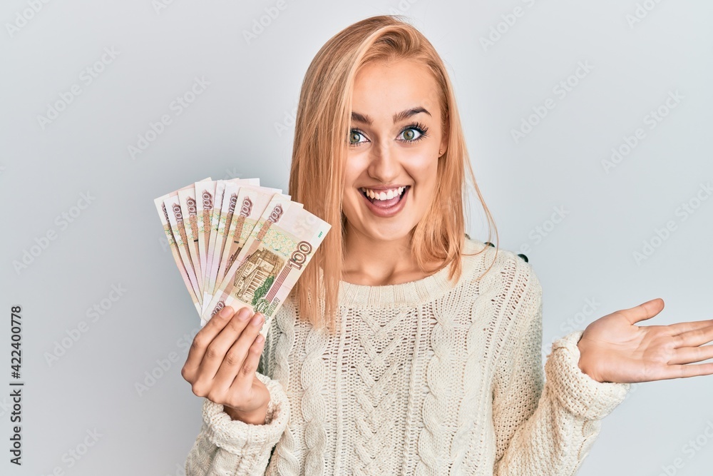 Beautiful caucasian woman holding 100 russian ruble banknotes celebrating achievement with happy smile and winner expression with raised hand Beautiful caucasian woman holding 100 russian ruble banknotes celebrating achievement with happy smile and winner expression with raised hand