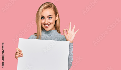 Beautiful caucasian woman holding blank empty banner doing ok sign with fingers, smiling friendly gesturing excellent symbol