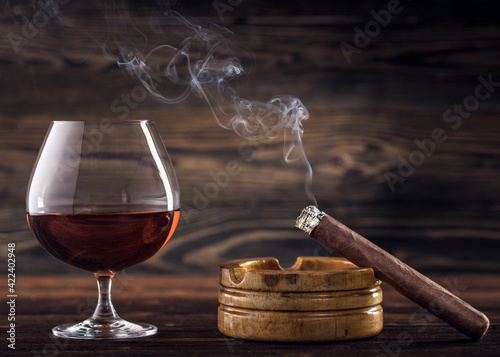 Cognac and cigar. Glass of cognac or brandy with cigars from Cuba Havana and ashtray on natural wooden background. Glass of whiskey with smoke cigar. Alcohol drink on Bar counter in the restaurant.   © artiom.photo