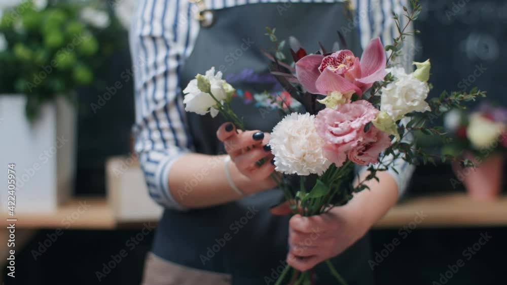 Handheld caucasian female florist makes beautiful bouquet, takes filler greenery and colorful flowers working at workplace in flower shop. Professional makes fresh bouquet for sale in floral store