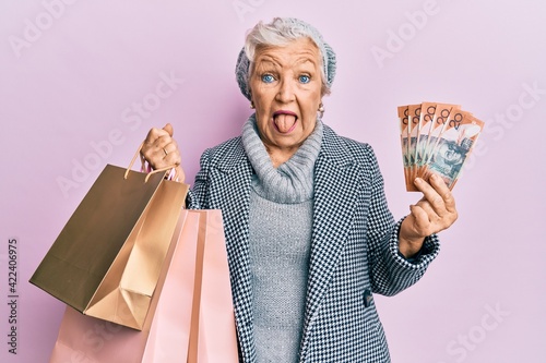 Senior grey-haired woman holding shopping bags and australia dollars sticking tongue out happy with funny expression.