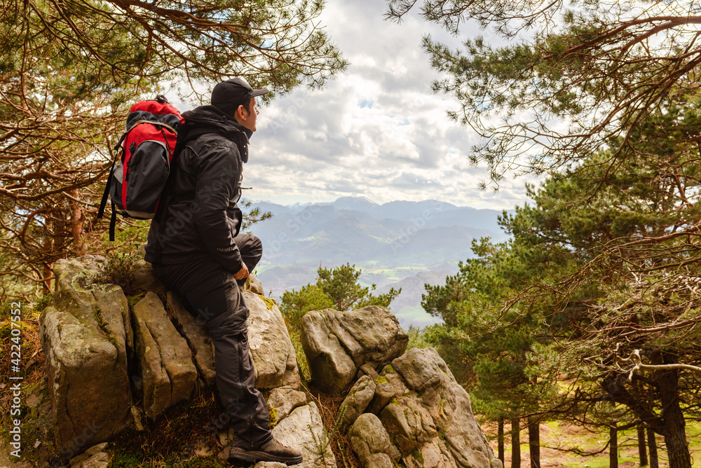 Naklejka premium hiker taking a break on his ascent to the mountain. Person observing the landscape from a forest. tired mountaineer, sitting on a rock looking at the mountains.