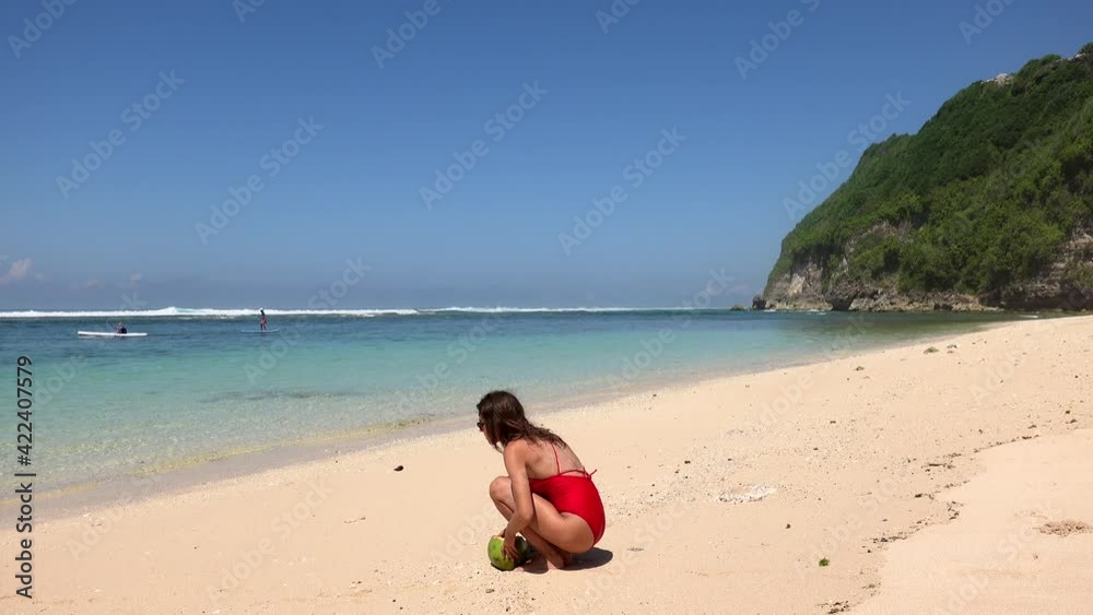 Slim tanned young woman in red bikini come and sitting on sand seashore and drinking coconut juice, enjoying wild beach in Bali, Indonesia Slim tanned young woman in red bikini come and sitting on sand seashore and drinking coconut juice, enjoying wild beach in Bali, Indonesia