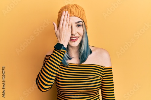 Young modern girl wearing wool hat covering one eye with hand, confident smile on face and surprise emotion.