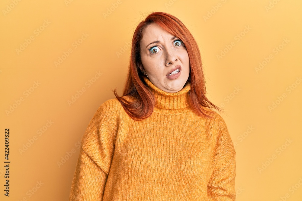 Beautiful redhead woman wearing casual winter sweater over yellow background in shock face, looking skeptical and sarcastic, surprised with open mouth Beautiful redhead woman wearing casual winter sweater over yellow background in shock face, looking skeptical and sarcastic, surprised with open mouth