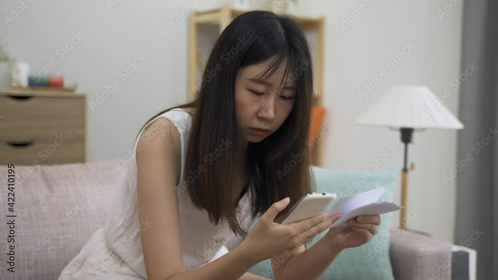 portrait agitated asian woman looking at her smaprtphone is checking lottery numbers one by one on the living room at home. portrait agitated asian woman looking at her smaprtphone is checking lottery numbers one by one on the living room at home.