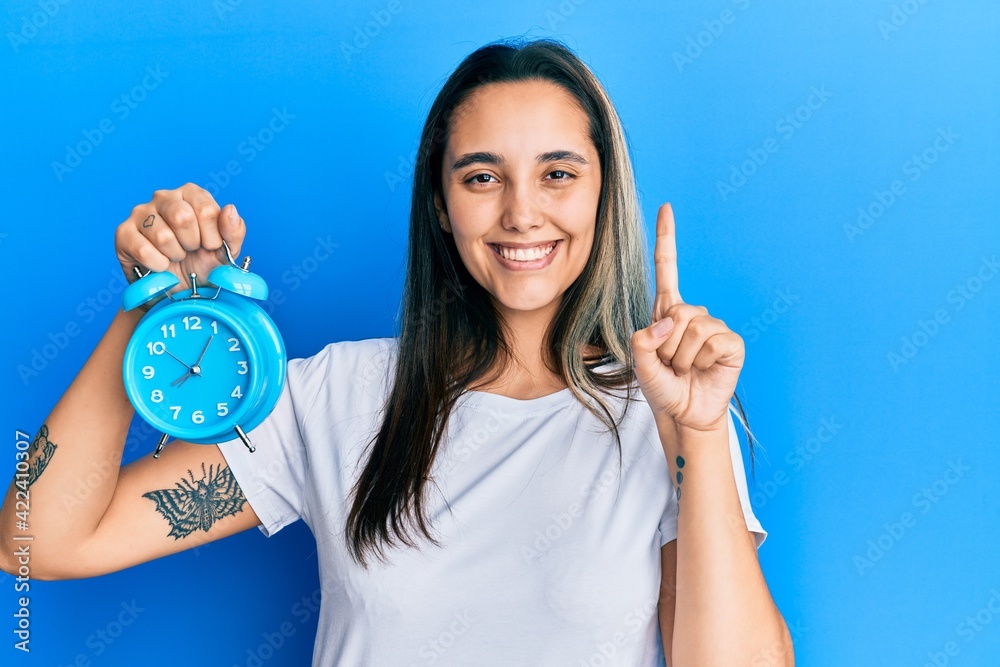 Young hispanic woman holding alarm clock smiling with an idea or question pointing finger with happy face, number one Young hispanic woman holding alarm clock smiling with an idea or question pointing finger with happy face, number one