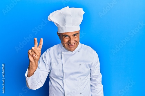 Mature middle east man wearing professional cook uniform and hat showing and pointing up with fingers number two while smiling confident and happy.