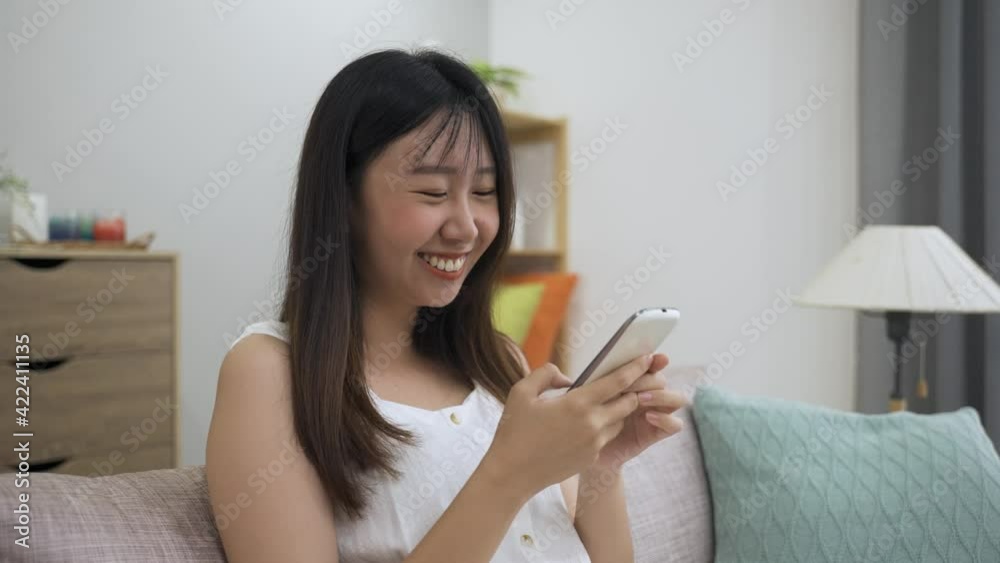 portrait beautiful asian girl is chuckling while enjoying funny social media feeds on her smartphone in the bright living room at home. portrait beautiful asian girl is chuckling while enjoying funny social media feeds on her smartphone in the bright living room at home.