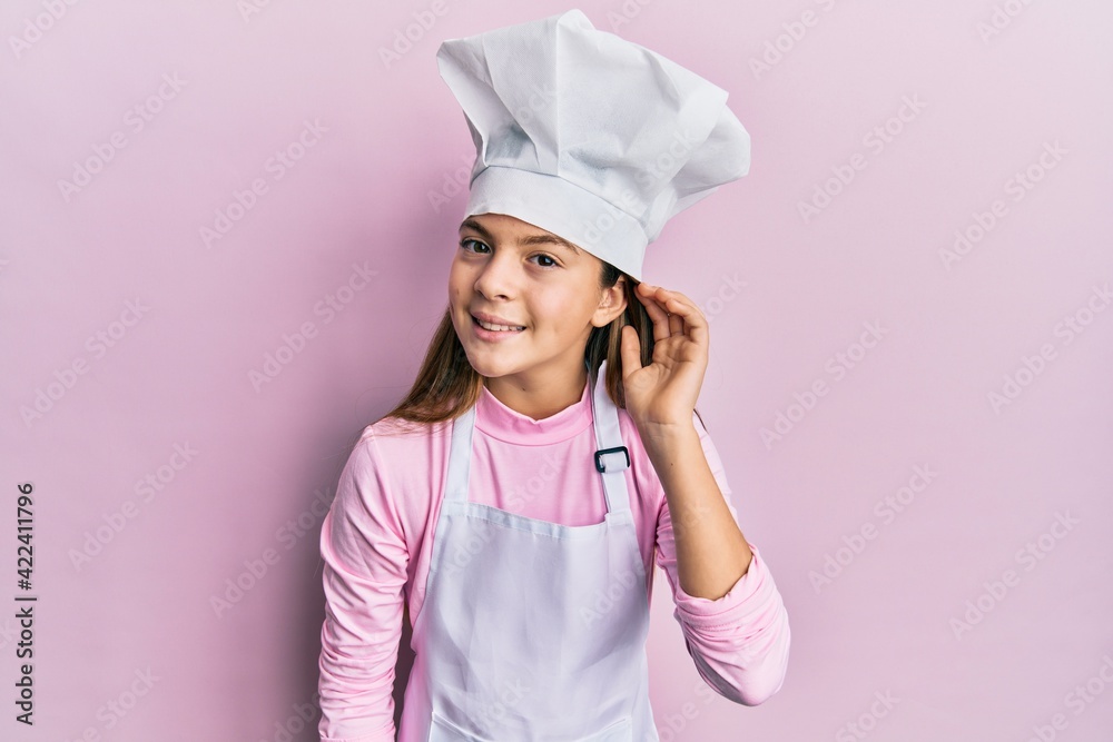 Beautiful brunette little girl wearing professional cook apron and hat smiling with hand over ear listening and hearing to rumor or gossip. deafness concept.