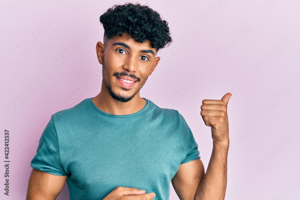 Young arab handsome man wearing casual clothes smiling with happy face looking and pointing to the side with thumb up. Young arab handsome man wearing casual clothes smiling with happy face looking and pointing to the side with thumb up.