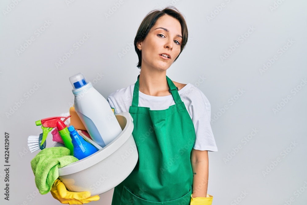 Young brunette woman with short hair wearing apron holding cleaning products looking sleepy and tired, exhausted for fatigue and hangover, lazy eyes in the morning. Young brunette woman with short hair wearing apron holding cleaning products looking sleepy and tired, exhausted for fatigue and hangover, lazy eyes in the morning.