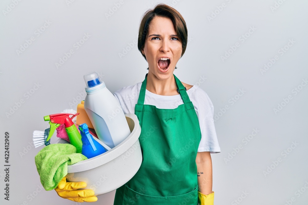 Young brunette woman with short hair wearing apron holding cleaning products angry and mad screaming frustrated and furious, shouting with anger. rage and aggressive concept. Young brunette woman with short hair wearing apron holding cleaning products angry and mad screaming frustrated and furious, shouting with anger. rage and aggressive concept.