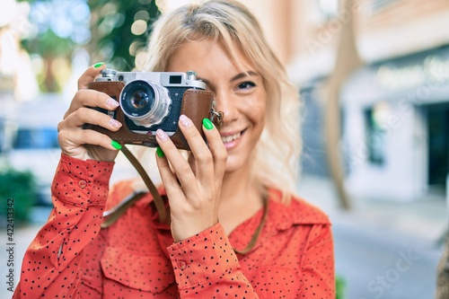 Young blonde woman smiling happy using vintage camera walking at the city.