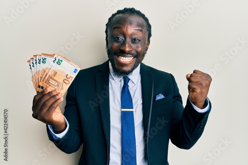 Handsome young black man wearing business suit holding 50 euros banknotes screaming proud, celebrating victory and success very excited with raised arm