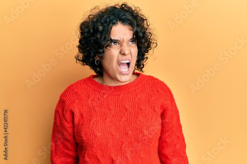 Young hispanic woman with curly hair wearing casual winter sweater angry and mad screaming frustrated and furious, shouting with anger. rage and aggressive concept. © Krakenimages.com