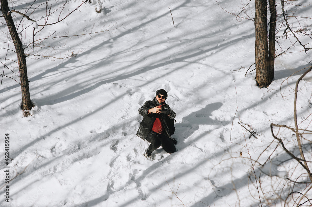 A wanderer, a tourist with a large black backpack on his back, travels through the snowy forest in different countries and takes pictures of the beautiful nature and himself on a mobile phone.
