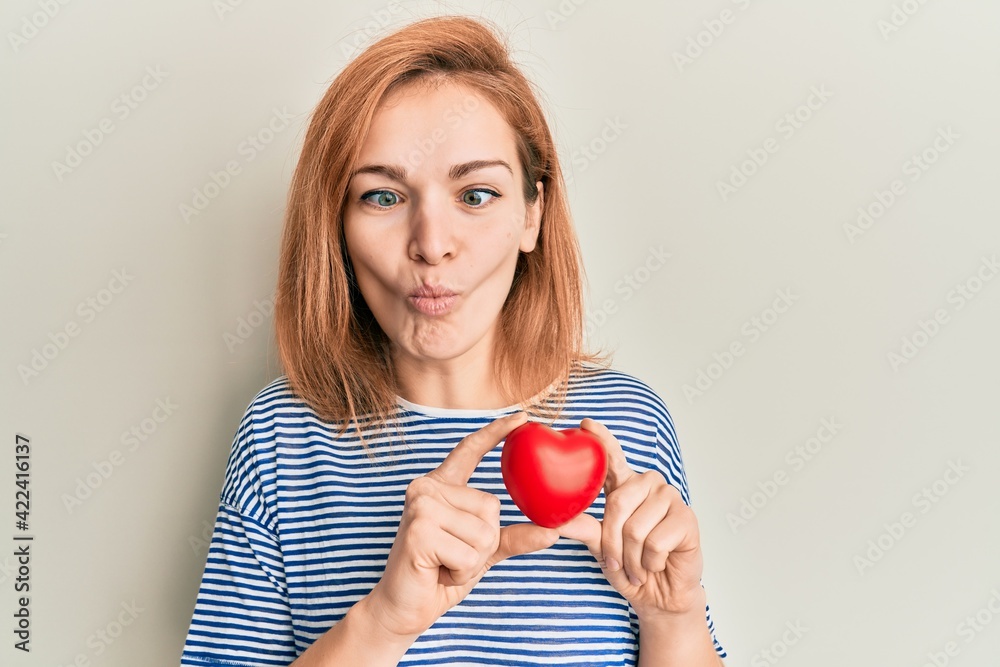 Young caucasian woman holding heart making fish face with mouth and squinting eyes, crazy and comical. Young caucasian woman holding heart making fish face with mouth and squinting eyes, crazy and comical.
